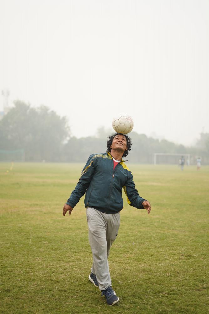 Male balancing football on head facing camera in a field. Wearing green zip up jacket and light grey jogging bottoms.