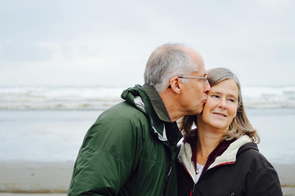 Male with glasses and short grey hair kissing woman on side of head, beach and sea in background