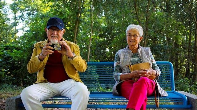 Older male sitting on blue bench holding a camera next to an older female in pink trousers. Both smiling.