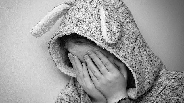 Black and white photo of young person with hands over face wearing a hooded top with rabbit ears on.