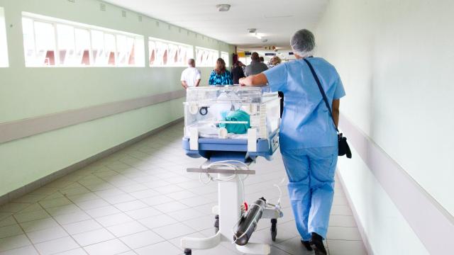 Photo of hospital corridor with staff member pushing incubator with young patient in and people walking away from camera in distance