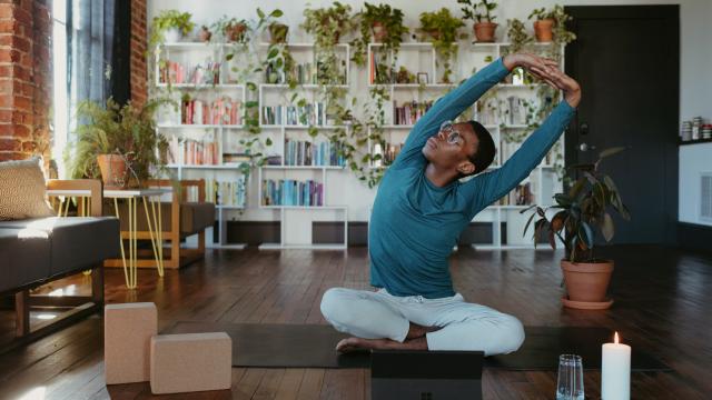 Person sat on yoga mat stretching with lit candle, glass of water and yoga blocks in front. Plant display shelf in background
