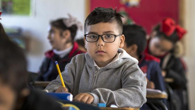 Boy in classroom sat at desk facing camera (wearing glasses) surrounded by other children out of focus