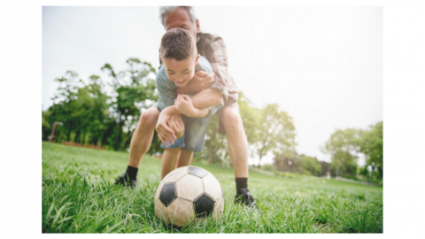 Image of young boy trying to get football on grassed field with sun shining whilst being embraced by older man with face obscured (depicting grandfather figure).