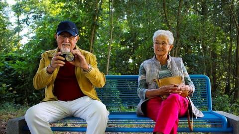 Older male sitting on blue bench holding a camera next to an older female in pink trousers. Both smiling.