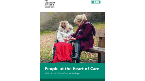 Female in mobility chair smiling at a lady sitting on a bench and holding hands in a green space with fallen leaves.