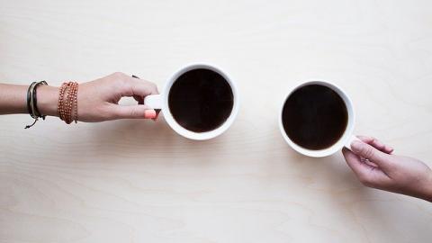 Two arms holding cups of black coffee on wooden table