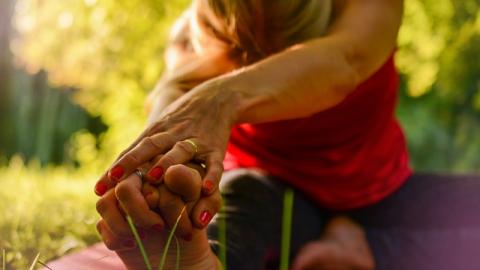 Lady sat on pink mat stretching arms to two with head down in outside green space