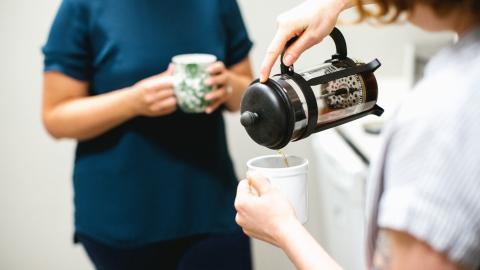 Person pouring coffee from cafetiere in white t-shirt at right hand side, person holding a cup in teal t-shirt in background left