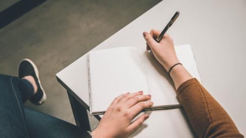Person sat at a table writing in open notebook wearing mustard top, dark blue jeans and black shoes