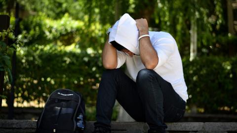 Young man sat down outside with head in hands, wearing white hooded top and dark jeans with black rucksack to left of him