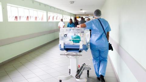 Photo of hospital corridor with staff member pushing incubator with young patient in and people walking away from camera in distance