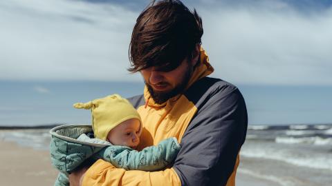 Photo of man in yellow and grey jacket holding young child wearing yellow wool hat on beach